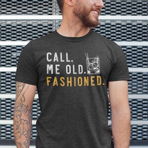 May include: A dark heather grey t-shirt with the text "CALL ME OLD. FASHIONED." in white with a graphic of an old fashioned cocktail in yellow.