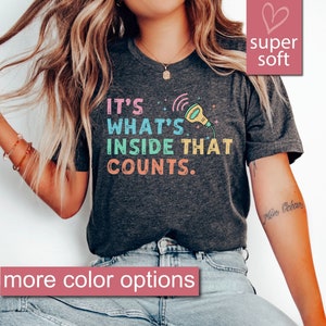 Op de afbeelding: Een donkergrijs heather t-shirt met de tekst "IT'S WHAT'S INSIDE THAT COUNTS." in regenboogkleuren. Het shirt heeft een cartoonillustratie van een microfoon met een regenboogkoord.
