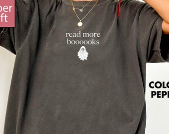 Può includere: T-shirt color pepe con la scritta "read more boooooks" e una grafica a forma di fantasma. La maglietta ha una vestibilità ampia e maniche corte. L'immagine include anche il testo "super soft" e "more color options".