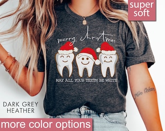 Puede incluir: Una camiseta de color gris oscuro jaspeado con un diseño navideño que presenta tres dientes con gorros de Papá Noel. El texto dice "merry christmas" y "MAY ALL YOUR TEETH BE WHITE".