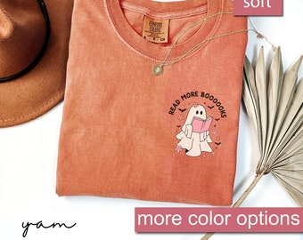 Può includere: Una maglietta color pesca con un'immagine di un fantasma che legge un libro con il testo "Read More Booooks".