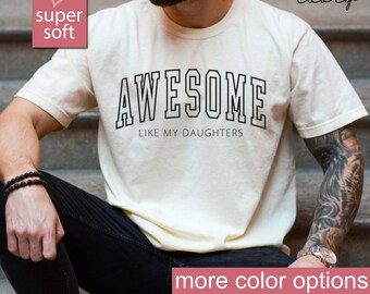 May include: A white t-shirt with black text that reads "AWESOME LIKE MY DAUGHTERS". The shirt is being worn by a person with tattoos on their arm.