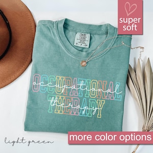 以下が含まれることがあります： 「OCCUPATIONAL THERAPY」の文字がカラフルなブロック体でプリントされた、ライトグリーンのTシャツ。ゴールドのネックレスと茶色の帽子も写っています。Tシャツには「super soft」と「more color options」と書かれています。