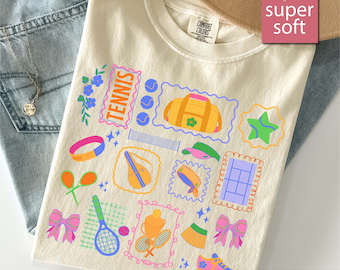 May include: Ivory t-shirt featuring a tennis-themed design with tennis balls, rackets, and a tennis bag. The word "TENNIS" is in a pink frame. The shirt is displayed on a pair of blue jeans.