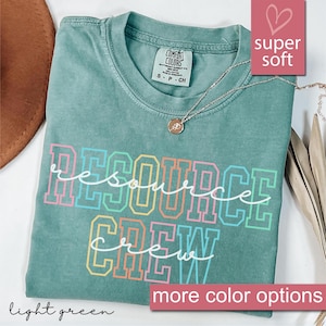 May include: A light green t-shirt with the words "RESOURCE CREW" in a colorful, block letter design. The shirt has a crew neck and short sleeves. A silver necklace with a pendant is draped on the shirt.