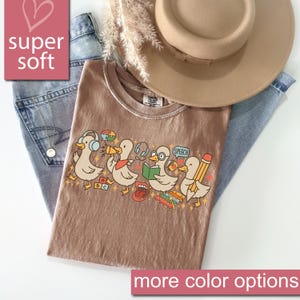 May include: A brown t-shirt with a graphic of cartoon ducks, each with a different accessory, such as headphones and a pencil. The shirt is paired with blue jeans and a tan hat. Text on the shirt includes "SPEECH" and "ABC". The words "super soft" and "more color options" are also visible.