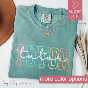 May include: Light green t-shirt with the word "tutor" in rainbow colored letters. The shirt is made of a soft, comfortable fabric.