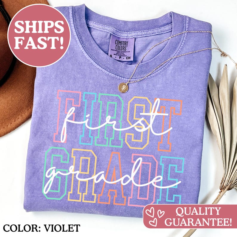 May include: A violet Comfort Colors t-shirt with the words "first grade" in a colorful, outlined font. A gold necklace with a pendant rests on the shirt. The shirt has a crew neck and is a solid color. The text "COLOR: VIOLET" is at the bottom.