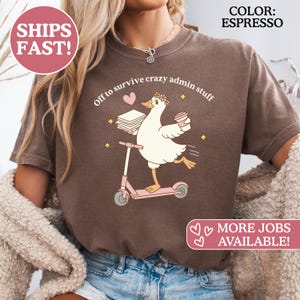 Pode incluir: Camiseta cor espresso com uma ilustração divertida de um ganso numa trotinete rosa, carregando papéis e uma chávena de café. O texto "Off to survive crazy admin stuff" está acima do ganso. Também tem o texto "MORE JOBS AVAILABLE!"