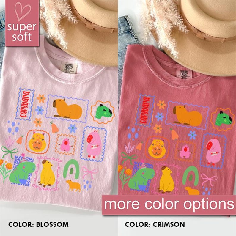 May include: Two t-shirts, one blossom pink and one crimson, feature a capybara graphic. The design includes capybara illustrations within postage stamp frames, along with floral and other decorative elements. The text "super soft" and "more color options" are also visible.