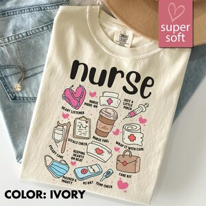 May include: Ivory-colored t-shirt with the word "nurse" in large black letters. The shirt features various medical-themed illustrations, including a stethoscope, coffee cup, and syringe. The text "super soft" is in a pink box.