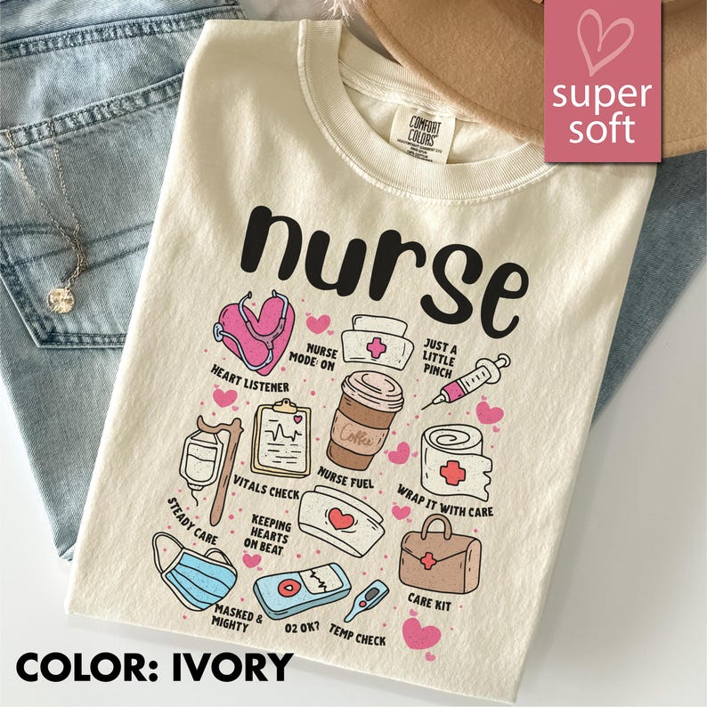 May include: Ivory-colored t-shirt with the word "nurse" in large black letters. The shirt features various medical-themed illustrations, including a stethoscope, coffee cup, and syringe. The text "super soft" is in a pink box.