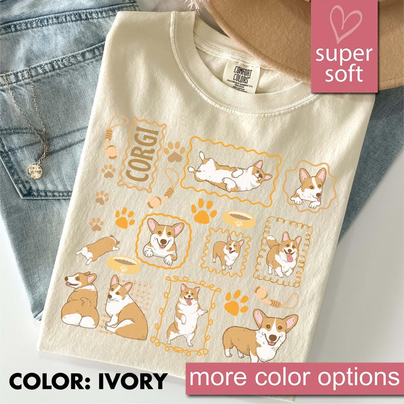 May include: Ivory-colored t-shirt featuring multiple illustrations of Corgi dogs in various poses, along with paw prints and food bowls. The word "CORGI" is printed on the shirt. The shirt is labeled "super soft" and "Comfort Colors".