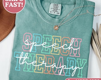 May include: A light green short-sleeve t-shirt featuring the text SPEECH THERAPY in a multi-colored block font with a white cursive script overlay. The shirt has a Comfort Colors brand label at the inner neckline and a thin gold-toned necklace.