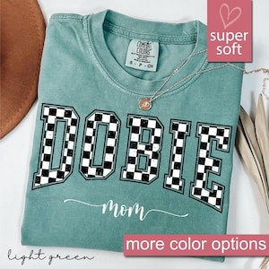 May include: Light green t-shirt with a checkered "DOBIE" graphic and the word "mom" written in cursive underneath. The shirt is made of a soft, comfortable fabric.