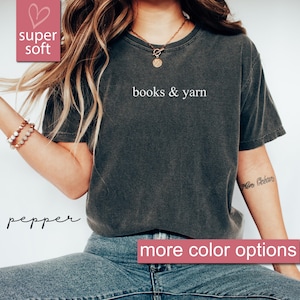 May include: A dark gray t-shirt with the words "books & yarn" in white. The shirt has a vintage, worn look. The text "super soft" is in a pink box in the upper left corner, and "more color options" is in a pink box at the bottom.