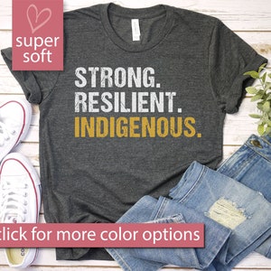 May include: A dark gray t-shirt with the words "STRONG. RESILIENT. INDIGENOUS." printed in white and gold lettering. The shirt is laid flat on a wooden surface with a pair of white sneakers and a pair of blue jeans.