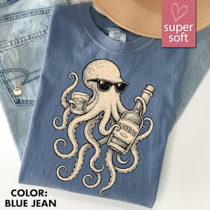 May include: Blue jean Comfort Colors t-shirt featuring a cartoon octopus wearing sunglasses and holding a glass of whiskey and a bourbon bottle. The shirt has the text "super soft" and "COLOR: BLUE JEAN".