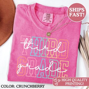Puede incluir: Una camiseta rosa Comfort Colors con las palabras "Third Grade" en un dise&ntilde;o colorido. La camiseta tiene cuello redondo y mangas cortas. Un collar de oro con un colgante est&aacute; drapeado sobre la camiseta.