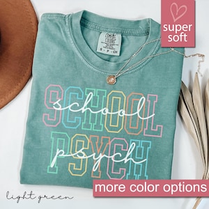 Op de afbeelding: Lichtgroen T-shirt met de tekst "school psych" in regenboogkleuren. De tekst is wit omrand. Het shirt is gemaakt van een zachte, comfortabele stof.