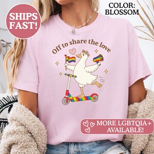 May include: Blossom-colored t-shirt with a whimsical graphic of a goose riding a rainbow scooter, holding a rainbow flag and a megaphone. The text "Off to share the love" is arched above the graphic. Additional text reads "MORE LGBTQIA+ AVAILABLE!"