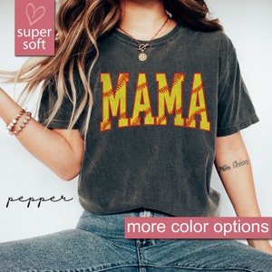 May include: A gray t-shirt with the word "MAMA" printed in yellow with a red outline. The letters are decorated with a softball stitch pattern. The shirt is being worn by a person with long blonde hair.