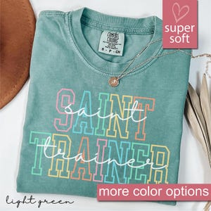 May include: A light green t-shirt with the text "Saint Trainer" in rainbow colors. The shirt is made of a soft, comfortable fabric and has a relaxed fit.