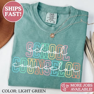 May include: A light green short-sleeve t-shirt with a crew neckline and a Comfort Colors brand label. The front features the text School Counselor in a layered design with block letters in pink, blue, and yellow, overlaid with white cursive script.