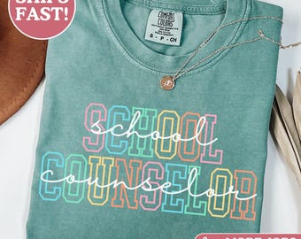 May include: A light green short-sleeve t-shirt with a crew neckline and a Comfort Colors brand label. The front features the text School Counselor in a layered design with block letters in pink, blue, and yellow, overlaid with white cursive script.