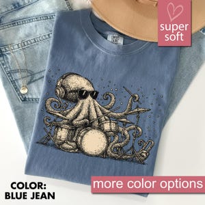 May include: Blue jean coloured t-shirt with an octopus illustration playing drums, wearing headphones and sunglasses. The text "COLOR: BLUE JEAN" is at the bottom left. The words "super soft" are in a pink heart-shaped box.