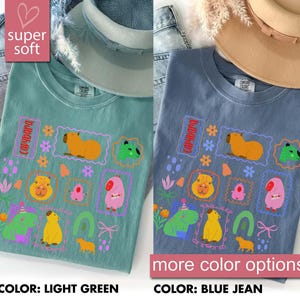 May include: Two t-shirts, one light green and one blue jean, featuring a colorful capybara graphic. The design includes various capybara illustrations within decorative frames, flowers, and the word "capybara". The shirts are labeled "super soft" and "more color options".