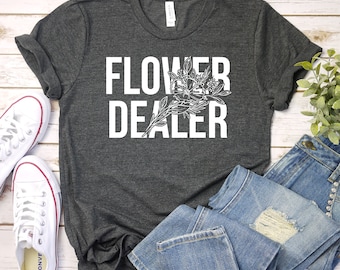 Op de afbeelding: Een donkergrijs T-shirt met de tekst "FLOWER DEALER" in witte letters. Een gestileerde bloemgrafiek is opgenomen in de tekst. Het shirt wordt getoond op een houten oppervlak met een paar witte sneakers en een blauwe jeans.