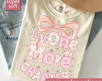 Op de afbeelding: Ivoor kleurig T-shirt met de tekst "Just One More Chapter" in roze en bloemmotieven. Het ontwerp bevat een grote roze strik en steraccenten. Het shirt wordt getoond met een hoed en jeans.