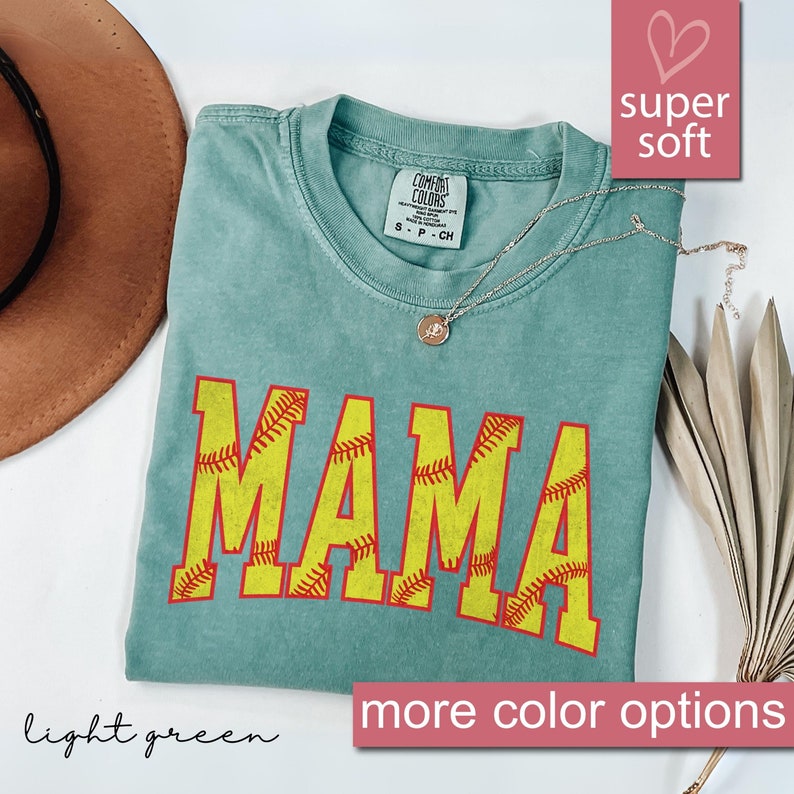 May include: A light green t-shirt with the word "MAMA" printed in yellow with red stitching, resembling a softball. The shirt is made of a soft, comfortable fabric.