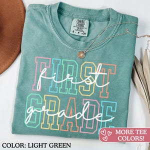 May include: Light green t-shirt with the words "First Grade" in a colorful, collegiate-style font. The shirt has a crew neck and short sleeves. A silver necklace with a small pendant is draped on the shirt.
