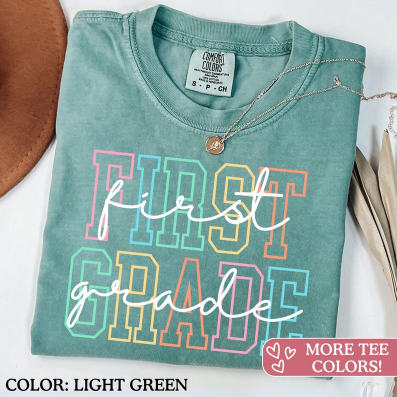May include: Light green t-shirt with the words "First Grade" in a colorful, collegiate-style font. The shirt has a crew neck and short sleeves. A silver necklace with a small pendant is draped on the shirt.