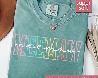 May include: A light green t-shirt with the word "MEEMAW" in colorful block letters. The shirt has a crew neck and short sleeves. A silver necklace with a pendant is draped on the shirt. The text "super soft" is in a pink box.