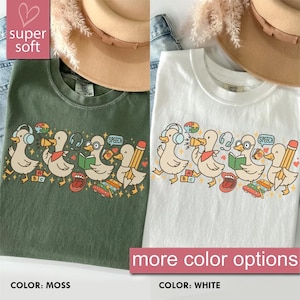 May include: Two t-shirts, one moss green and one white, featuring a cartoon duck design. The ducks are depicted with headphones, books, and pencils. The text "super soft" and "more color options" are also visible.