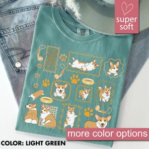 May include: Light green t-shirt with a graphic of multiple corgi dogs in various poses, surrounded by paw prints, dog bowls, and toys. The text "CORGI" is also present. The shirt is labeled "super soft" and "more color options".