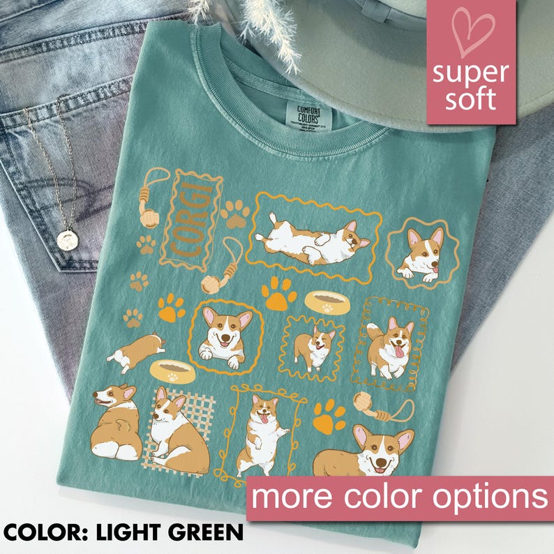 May include: Light green t-shirt with a graphic of multiple corgi dogs in various poses, surrounded by paw prints, dog bowls, and toys. The text "CORGI" is also present. The shirt is labeled "super soft" and "more color options".
