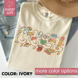 Puede incluir: Camiseta de color marfil con un diseño de patos de dibujos animados relacionados con la terapia del habla y la educación. Los patos se representan con auriculares, megáfonos, libros y lápices. El texto "COLOR: IVORY" y "more color options" es visible.