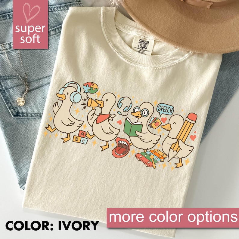 May include: Ivory-colored t-shirt featuring a playful design of cartoon ducks with speech therapy and educational elements. The ducks are depicted with headphones, megaphones, books, and pencils. The text "COLOR: IVORY" and "more color options" are visible.
