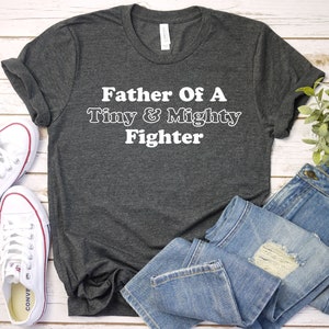 May include: A dark gray t-shirt with the text "Father Of A Tiny & Mighty Fighter" in white.