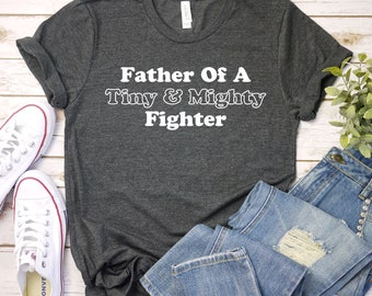 May include: A dark gray t-shirt with the text "Father Of A Tiny & Mighty Fighter" in white.