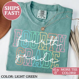 May include: Light green Comfort Colors t-shirt with the words "Fourth Grade" in a colourful, outlined font. A silver necklace with a pendant rests on the shirt. The shirt has a crew neck and short sleeves.
