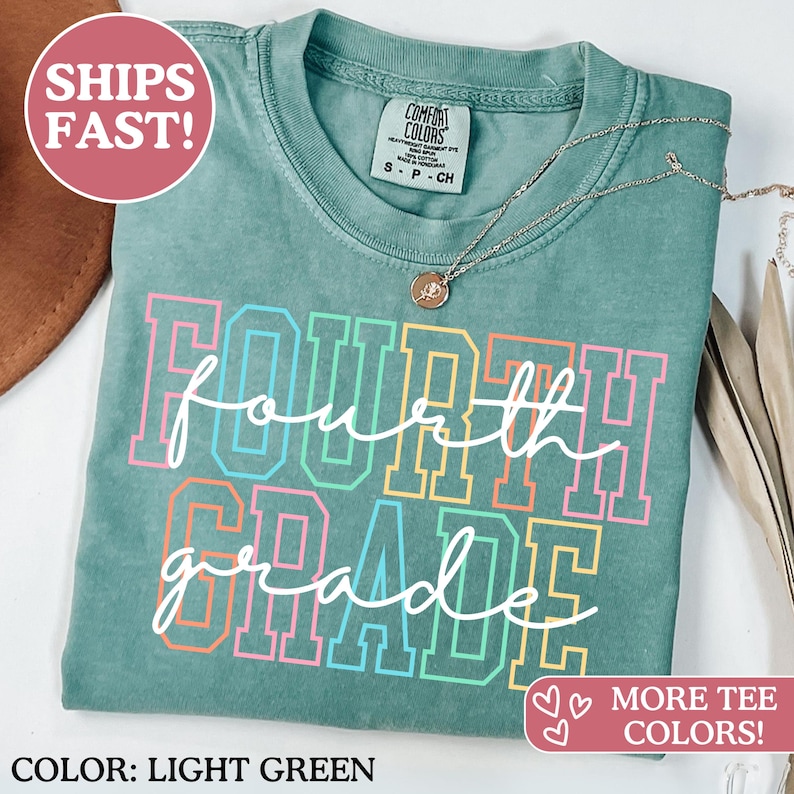 May include: Light green Comfort Colors t-shirt with the words "Fourth Grade" in a colourful, outlined font. A silver necklace with a pendant rests on the shirt. The shirt has a crew neck and short sleeves.