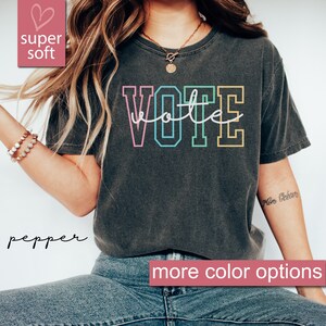 May include: A dark grey t-shirt with the word "VOTE" printed in rainbow colors. The word "vote" is written in cursive script inside the letters. The text "super soft" and "more color options" are also visible in the image.