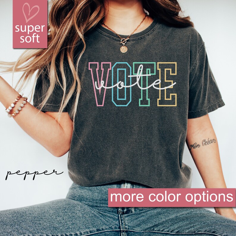 May include: A dark grey t-shirt with the word "VOTE" printed in rainbow colors. The word "vote" is written in cursive script inside the letters. The text "super soft" and "more color options" are also visible in the image.