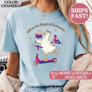 May include: Light blue t-shirt with the text "Off to be fluid & fantastic." The graphic features a cartoon goose on a scooter, holding a bisexual pride flag and gender symbols. The shirt is chambray in color.