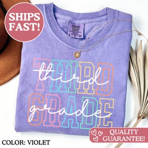 Puede incluir: Camiseta de color violeta con las palabras "third grade" en una fuente colorida y delineada. La camiseta tiene cuello redondo y un collar dorado. El texto "COLOR: VIOLET" est&aacute; en la parte inferior izquierda.